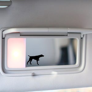 May include: A white car visor with a mirror and a light. A black silhouette of a dog is visible in the reflection of the mirror.