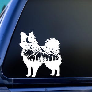 May include: White die-cut vinyl decal of a dog silhouette with a mountain and forest landscape design. The dog's body is filled with a night sky scene, including a crescent moon and stars. The decal is applied to a dark surface.