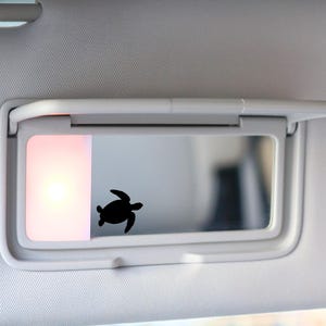 May include: A black turtle silhouette is affixed to the mirror of a car visor. The visor is white and has a built-in light. The light is on, emitting a soft, warm glow. The background is out of focus.