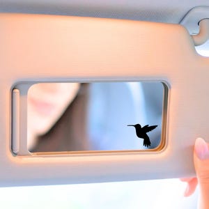 May include: A car visor with a rectangular mirror. A black hummingbird silhouette is affixed to the mirror. The visor is beige and is being held by a person with a pink manicure.