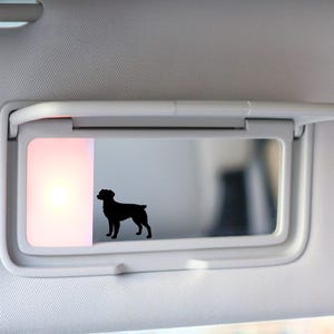 May include: A black silhouette of a dog is displayed on a car visor mirror. The visor is light gray, and the mirror is rectangular. The dog is facing right, with its tail slightly raised. The background is blurred.
