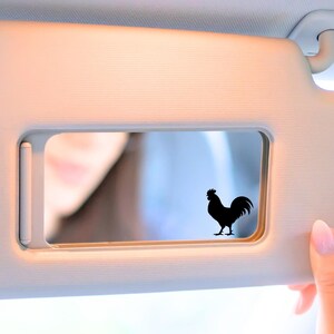 May include: A light-colored car visor with a rectangular mirror. A black silhouette of a rooster is affixed to the mirror. A person's hand with pink nail polish is holding the visor.