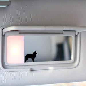 May include: A car visor with a built-in mirror. The mirror reflects a black silhouette of a dog. The visor is light gray, and the mirror has a rectangular light on the left side.