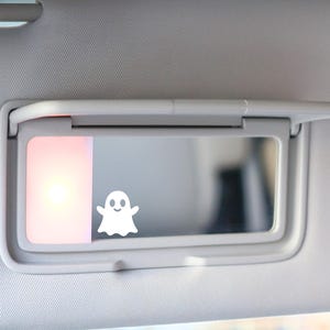 May include: A car visor with a built-in mirror and a small, white ghost sticker. The visor is gray and has a light illuminating the mirror. The ghost sticker has a friendly face.