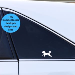May include: White silhouette of a dog running on a black car window. The window is tinted and the car is white. A blue circle with the text "Tiny Doodle Decals -Multiple designs and sizes" is in the upper left corner of the image.