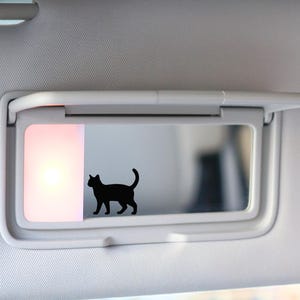 May include: A black cat silhouette decal is affixed to a car visor mirror. The visor is white and has a built-in light. The cat is walking with its tail curved upwards.