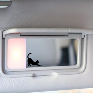 May include: A black cat stretching decal is affixed to a car visor mirror. The visor is white and has a built-in light. The background is out of focus, suggesting the interior of a vehicle.