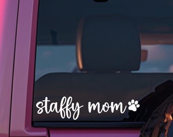 May include: White vinyl decal reading "staffy mom" with a paw print, affixed to a vehicle window. The text is in a cursive font. The vehicle's exterior is pink. The decal is designed for a dog owner.
