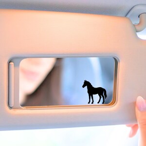 May include: A black horse silhouette sticker is affixed to a car visor. The visor is a light beige color, and a person's hand is visible, holding the visor. The background is blurred, showing a person's face.