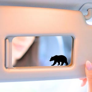 May include: A car sun visor with a black bear silhouette decal. The visor is a light beige color, and the bear is positioned in the center of the mirror. A hand with pink nail polish is holding the visor.
