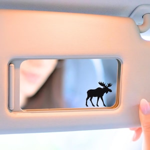 May include: A black moose silhouette sticker is affixed to a car visor. The visor is a light beige color, and a person's hand with pink nail polish is visible. The background shows a blurred reflection.
