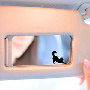 May include: A black cat silhouette sticker is affixed to a car visor mirror. The visor is a light beige color, and a person's hand with pink nail polish is visible. The cat is in a playful pose.