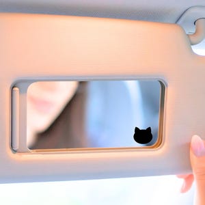 May include: A white car sun visor with a mirror and a black cat silhouette sticker. The mirror is open and shows a reflection of a person's face.