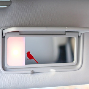 May include: A white car visor with a small mirror and a red cardinal silhouette sticker. The mirror has a light inside.