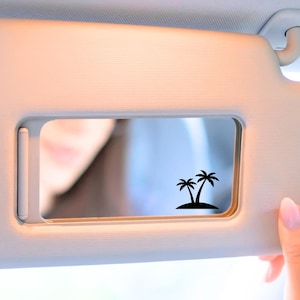 May include: A light beige car visor with a rectangular mirror. The mirror features a black silhouette of two palm trees on a small island. A hand with pink nail polish is holding the visor.