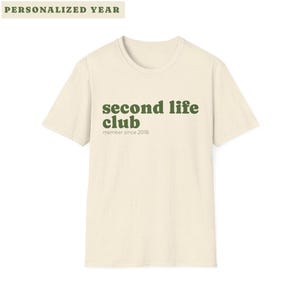 May include: A cream-coloured t-shirt featuring the words "second life club" in green, with "member since 2018" underneath. The shirt has a crew neck and short sleeves. The words "PERSONALIZED YEAR" are at the top.