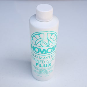 May include: A white bottle of Novacan Old Master Stained Glass Flux. The bottle has a turquoise label with the company logo and the product name.