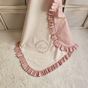 May include: A baby blanket in soft pink and cream colors, featuring a ruffled edge. The blanket has an embroidered floral wreath with the name "Lexi" in the center. The blanket is folded and displayed on a white surface.