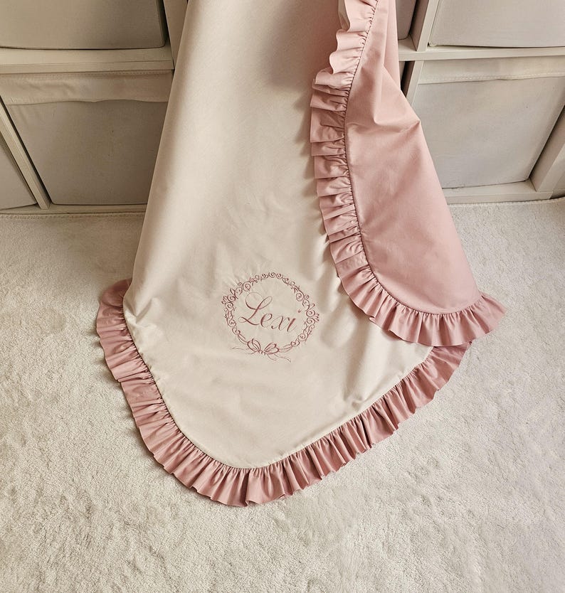 May include: A baby blanket in soft pink and cream colors, featuring a ruffled edge. The blanket has an embroidered floral wreath with the name "Lexi" in the center. The blanket is folded and displayed on a white surface.