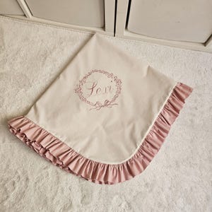May include: A light beige baby blanket with a pink ruffled edge. The blanket features the name "Lexi" embroidered in a floral wreath design. The blanket is folded on a white surface.