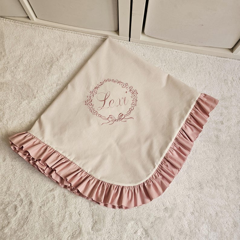 May include: A light beige baby blanket with a pink ruffled edge. The blanket features the name "Lexi" embroidered in a floral wreath design. The blanket is folded on a white surface.