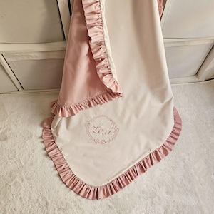 May include: A soft, two-tone baby blanket in pink and cream, trimmed with ruffled edges. The blanket features the name "Lexi" embroidered in a floral wreath design, adding a personalized touch to the nursery decor.