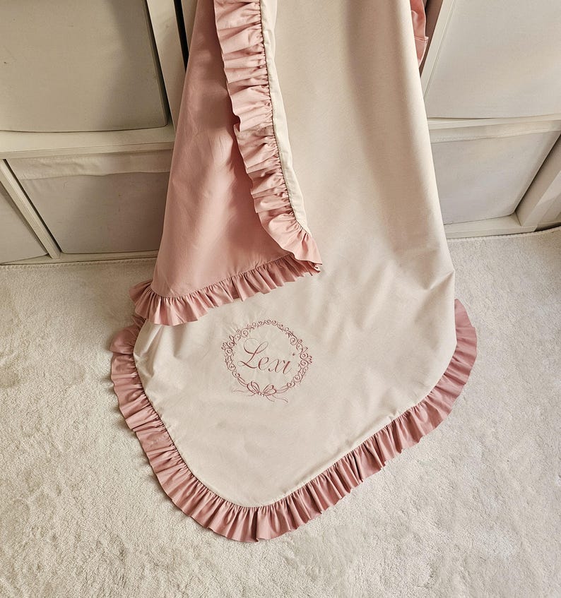 May include: A soft, two-tone baby blanket in pink and cream, trimmed with ruffled edges. The blanket features the name "Lexi" embroidered in a floral wreath design, adding a personalized touch to the nursery decor.