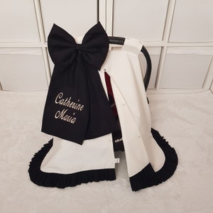 Könnte beinhalten: Weißer Kinderwagenbezug mit schwarzem Rüschenbesatz und einer großen schwarzen Schleife mit einem personalisierten Sticknamen "Catherine Maria" in goldfarbener Stickerei.