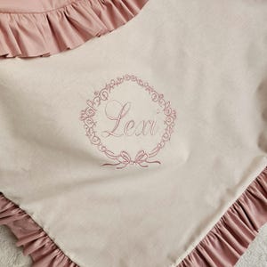 May include: A light pink baby blanket with a ruffled edge. The blanket features an embroidered wreath design with the name "Lexi" in the center. The blanket is made of a soft, light-colored fabric.