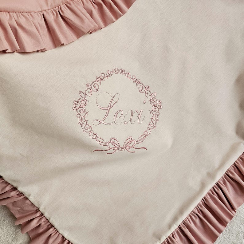 May include: A light pink baby blanket with a ruffled edge. The blanket features an embroidered wreath design with the name "Lexi" in the center. The blanket is made of a soft, light-colored fabric.