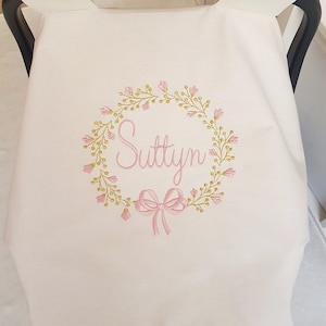 May include: A light pink baby car seat cover with an embroidered floral wreath design. The wreath surrounds the name "Suttyn" in pink script, with a pink bow at the bottom. The cover is made of soft fabric.
