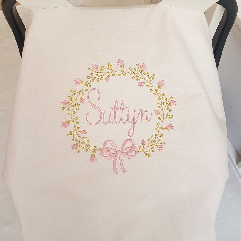 May include: A light pink baby car seat cover with an embroidered floral wreath design. The wreath surrounds the name "Suttyn" in pink script, with a pink bow at the bottom. The cover is made of soft fabric.