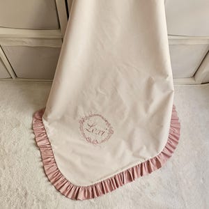 May include: A light pink baby blanket with a ruffled edge in a darker pink. The blanket has the name "Lexi" embroidered in a floral wreath design. The blanket is draped over a white surface.