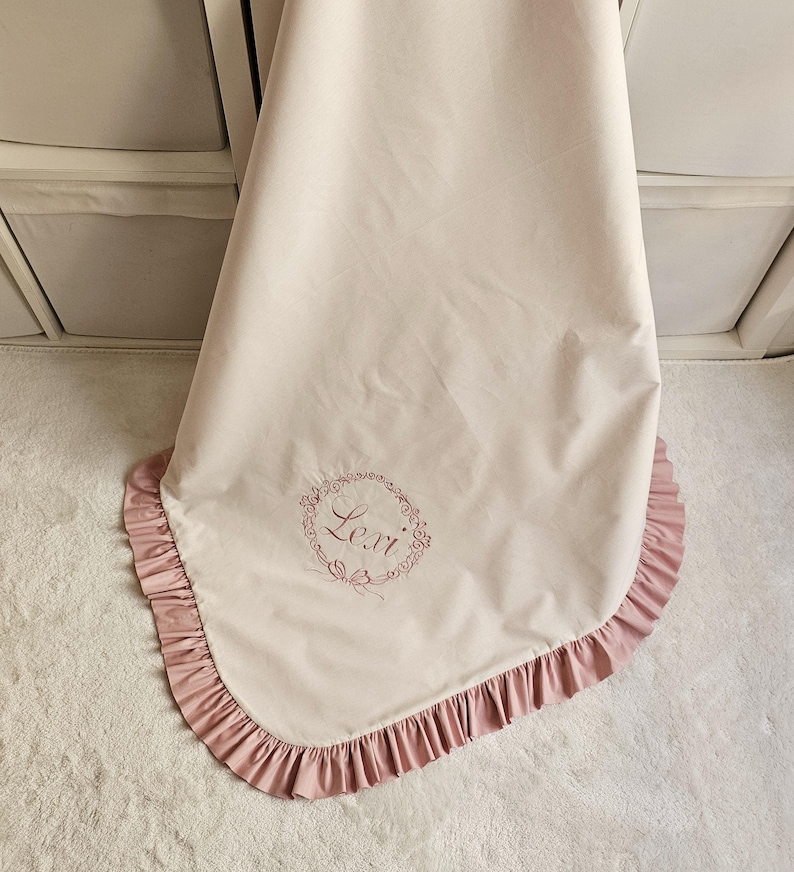 May include: A light pink baby blanket with a ruffled edge in a darker pink. The blanket has the name "Lexi" embroidered in a floral wreath design. The blanket is draped over a white surface.
