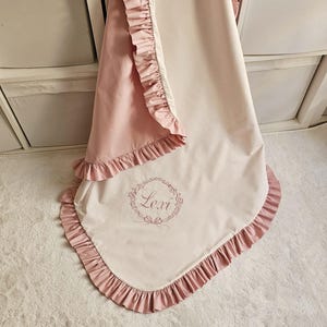 May include: A baby blanket in soft pink and cream colors, featuring a ruffled edge. The blanket has the name "Lexi" embroidered in a floral wreath design. The blanket is made of a soft, possibly satin-like material.