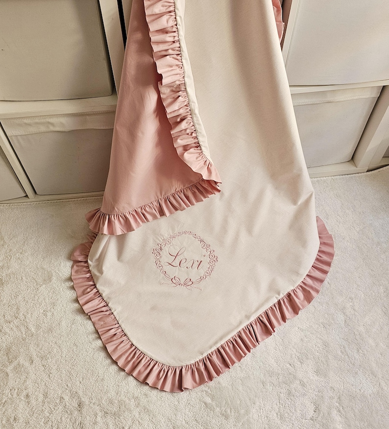 May include: A baby blanket in soft pink and cream colors, featuring a ruffled edge. The blanket has the name "Lexi" embroidered in a floral wreath design. The blanket is made of a soft, possibly satin-like material.