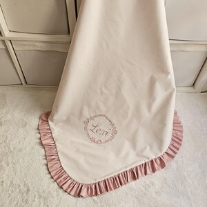 May include: A light pink baby blanket with a ruffled edge in a darker pink. The blanket has the name "Lexi" embroidered in a circular floral design. The blanket is made of a soft, smooth fabric.