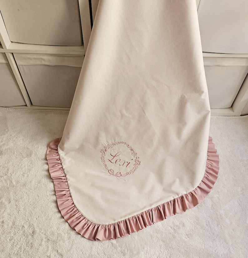 May include: A light pink baby blanket with a ruffled edge in a darker pink. The blanket has the name "Lexi" embroidered in a circular floral design. The blanket is made of a soft, smooth fabric.