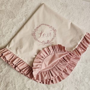 May include: A light beige baby blanket with a pink ruffled edge. The blanket has the name "Lexi" embroidered in a floral wreath design. The blanket is made of soft fabric.