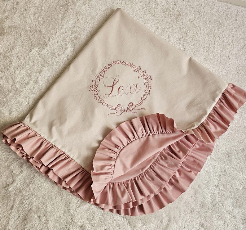 May include: A light beige baby blanket with a pink ruffled edge. The blanket has the name "Lexi" embroidered in a floral wreath design. The blanket is made of soft fabric.