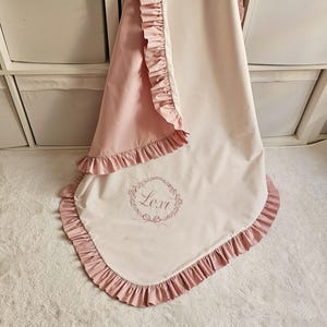 May include: A baby blanket with a pink ruffled trim and a light pink backing. The blanket has the name "Lexi" embroidered in a floral wreath design. The blanket is made of a soft, lightweight material.