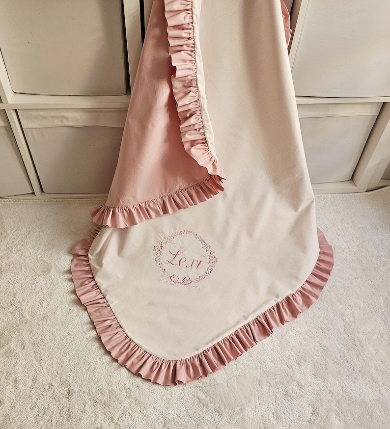 May include: A baby blanket with a pink ruffled trim and a light pink backing. The blanket has the name "Lexi" embroidered in a floral wreath design. The blanket is made of a soft, lightweight material.