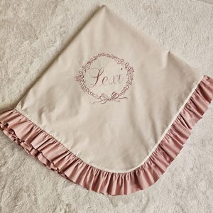 May include: A beige baby blanket with a pink ruffled edge. The blanket features the name "Lexi" embroidered in a floral wreath design. The blanket is made of soft fabric.