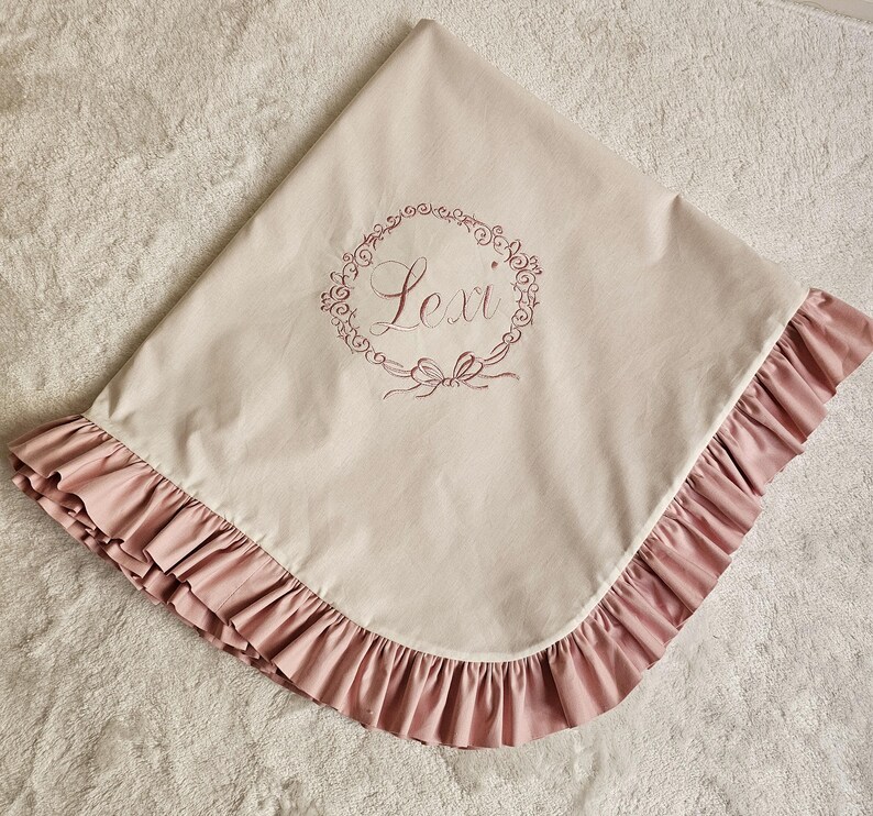 May include: A beige baby blanket with a pink ruffled edge. The blanket features the name "Lexi" embroidered in a floral wreath design. The blanket is made of soft fabric.