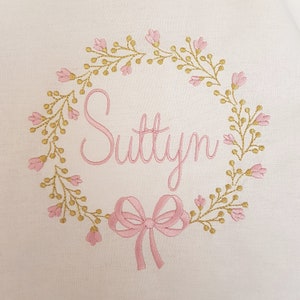 May include: Embroidered design on a white background. The design features a floral wreath in gold and pink, surrounding the name "Suttyn" in pink script. A pink bow is at the bottom of the design. The embroidery is detailed and delicate.