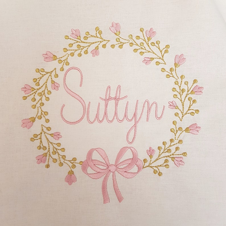 May include: Embroidered design on a white background. The design features a floral wreath in gold and pink, surrounding the name "Suttyn" in pink script. A pink bow is at the bottom of the design. The embroidery is detailed and delicate.