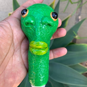 May include: Green ceramic pipe shaped like a cartoon alien with black eyes and a yellow mouth. The pipe has a textured surface.
