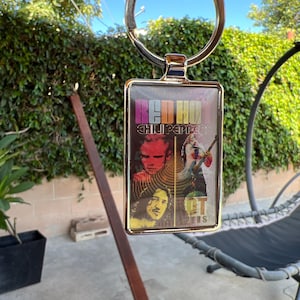 May include: A rectangular keyring with a silver-coloured metal frame. The keyring features a colourful graphic of the Red Hot Chili Peppers band members and the band's name. The background is a mix of red, yellow, and black.