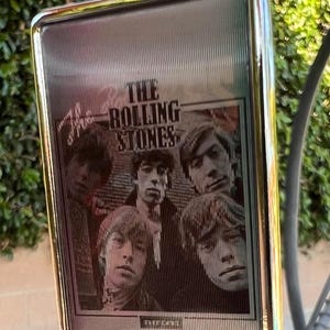 Può includere: Un portachiavi rettangolare con cornice argentata e anello argentato. Il portachiavi presenta un'immagine in bianco e nero dei membri della band The Rolling Stones. Il testo "THE ROLLING STONES" è visibile.