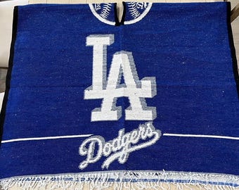 Op de afbeelding: Een blauwe poncho met het Los Angeles Dodgers-logo in wit. De poncho heeft een witte franje langs de onderrand en een witte streep in het midden. De letters "LA" staan boven het woord "Dodgers".